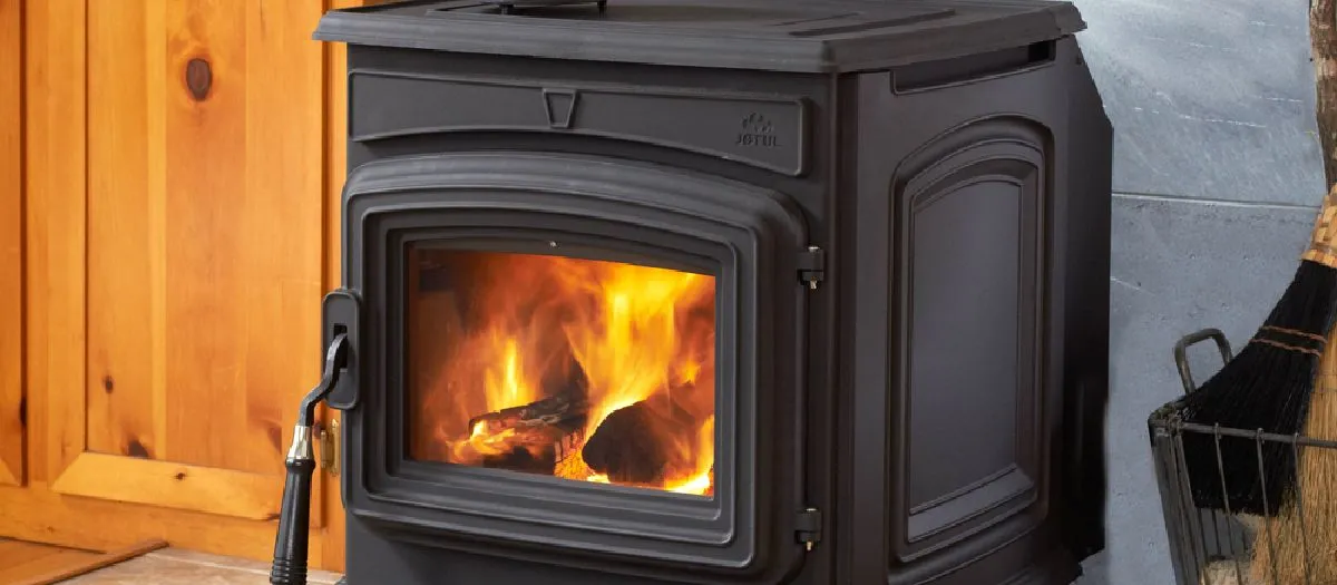 A black wood-burning stove with a glass door displays a brightly burning fire inside. The stove is set against wooden paneling and a stone wall, with a broom and basket nearby.