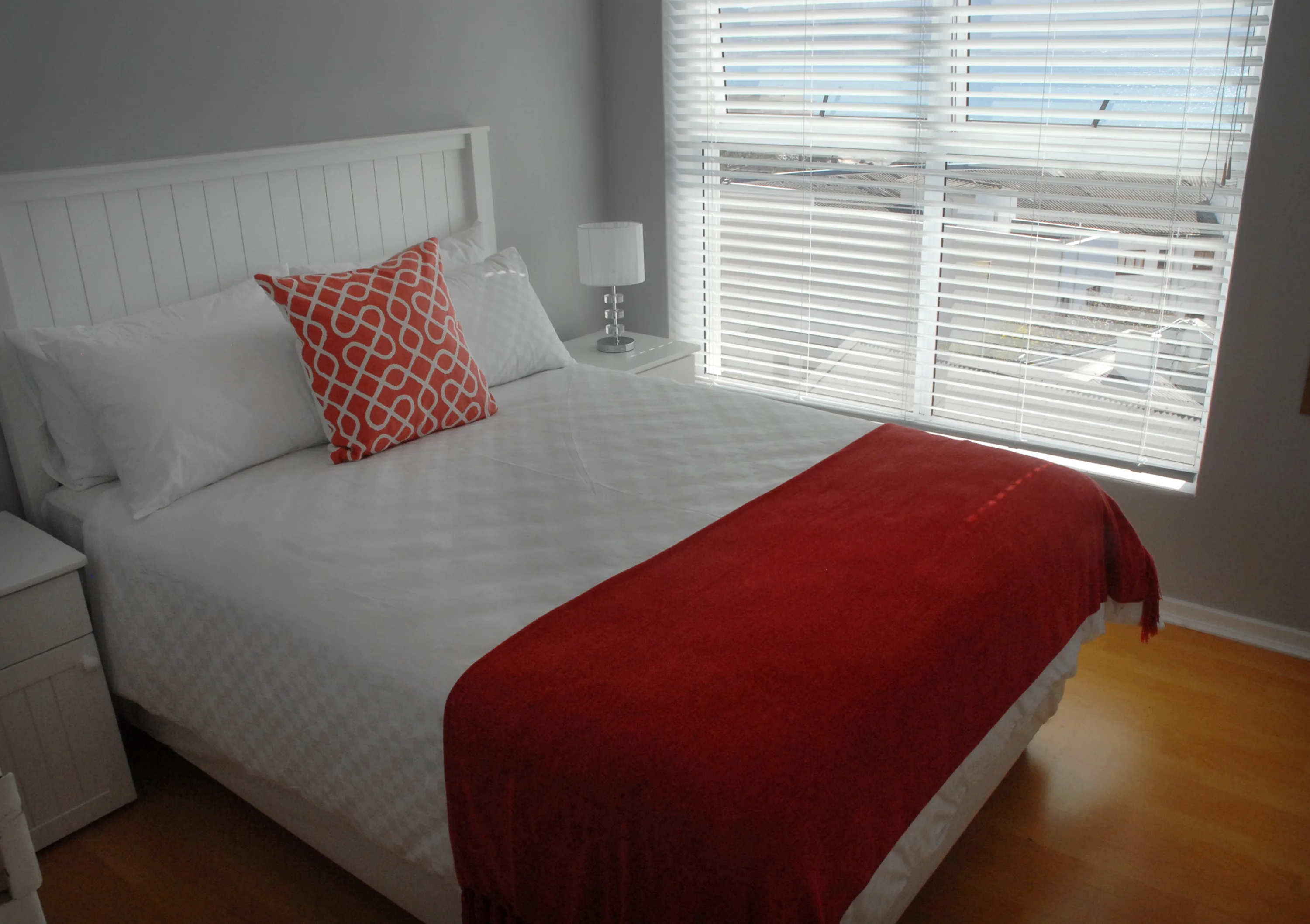 A cozy bedroom with a white bed, red and white decorative pillow, red blanket at the end of the bed, side table with a lamp, and blinds covering a large window.