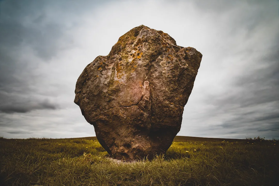 A large, weathered standing stone sits amid a grassy landscape under a cloudy, overcast sky. Small patches of yellow lichen are visible on the surface of the stone.