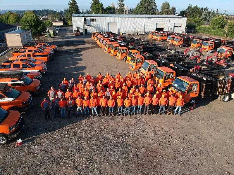 A large group of workers in orange safety gear and hard hats stand together in a lot with numerous orange trucks and work vehicles, near a large industrial building.