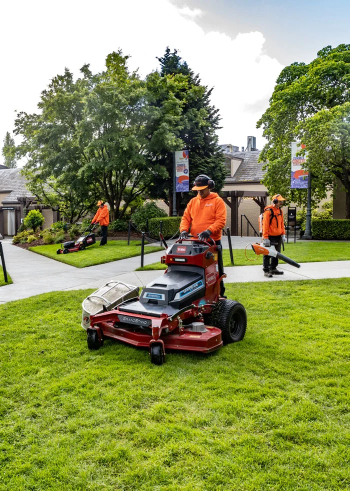 Three workers dressed in orange operate landscaping equipment, including a riding lawn mower, a push mower, and a leaf blower, on a well-maintained lawn with trees and a building in the background.