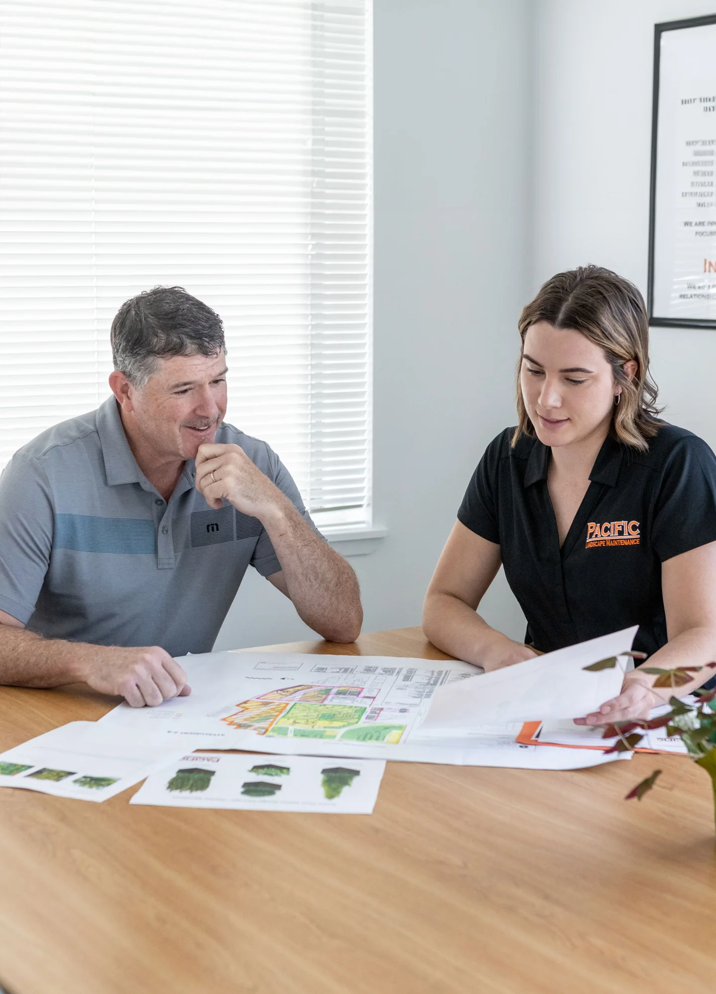A man and woman sit at a table discussing a landscape design plan. The woman is holding a document while the man looks on thoughtfully. Various papers and plant images are spread on the table.