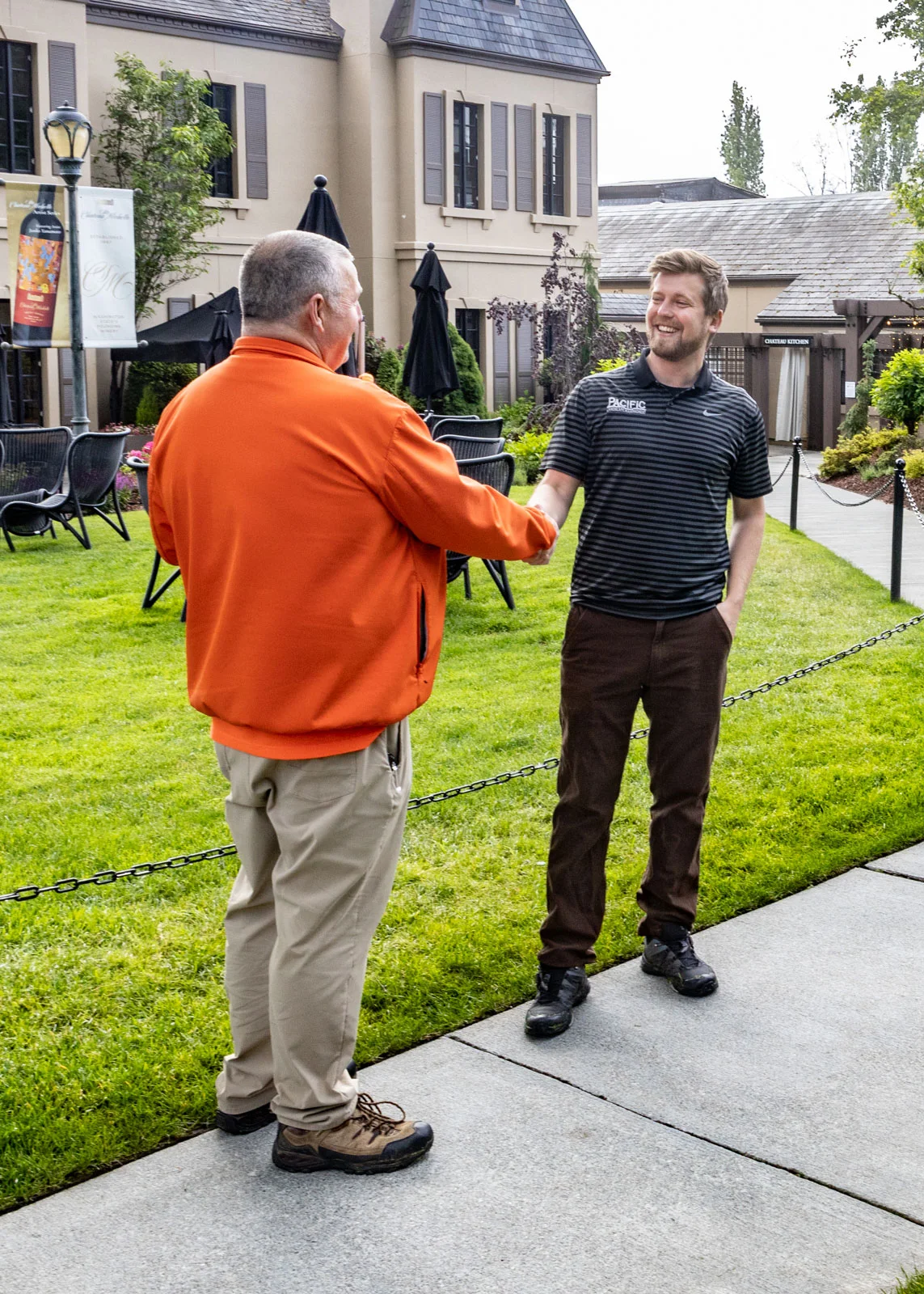 Two men are shaking hands outdoors in front of a beige building with multiple windows. One man wears an orange jacket and tan pants, while the other wears a striped shirt and brown pants.