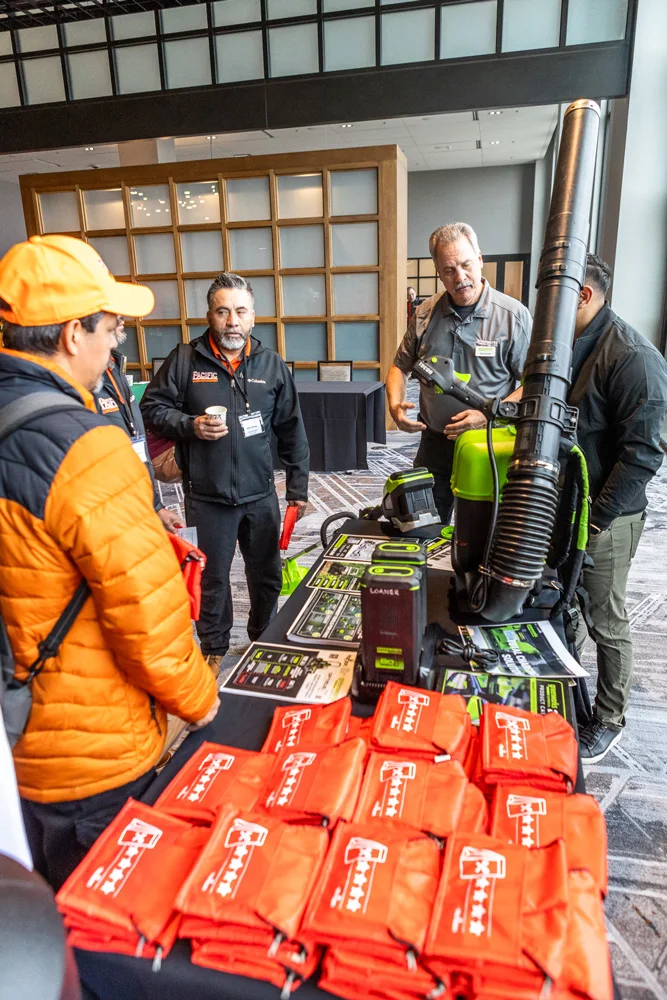 Four men are gathered around a table exhibiting leaf blowers and other equipment at a trade show. Orange bags with white logos are neatly stacked on the table.