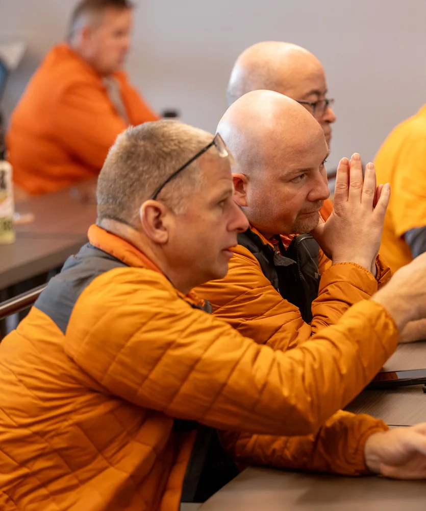 Men in orange jackets sit at a table in a classroom setting. One man gestures with his hands while speaking. Others are focused on the discussion, with laptops and water bottles on the table.