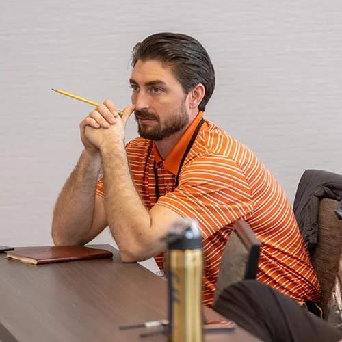 A man in an orange striped shirt sits at a desk with a notebook and pen, holding a pencil near his face, appearing deep in thought. A jacket and a water bottle are beside him on the desk.