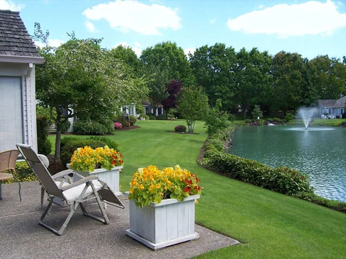 A backyard patio with two lounge chairs and flower pots, overlooking a lush green lawn with trees, shrubs, and a pond with a fountain under a partly cloudy sky.