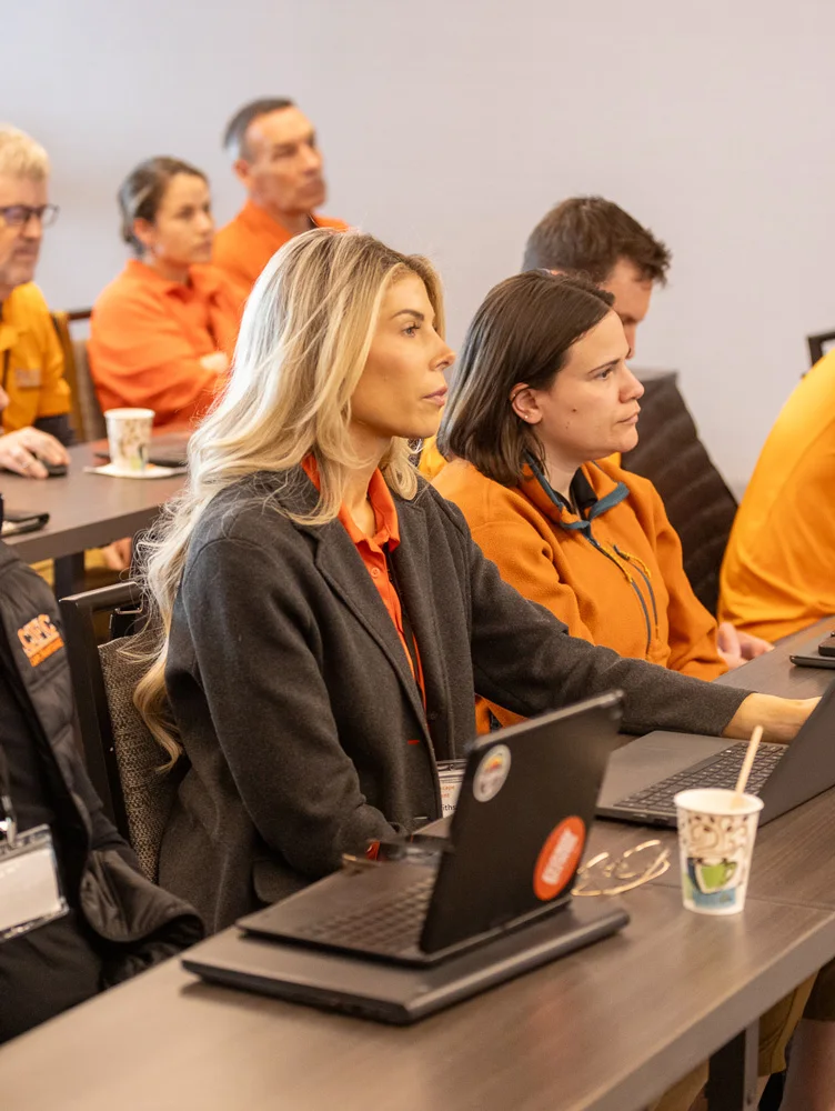 People seated at desks in a conference room, attentively looking ahead. Some are wearing orange shirts, while others have laptops and notepads. Coffee cups are placed on the desks.
