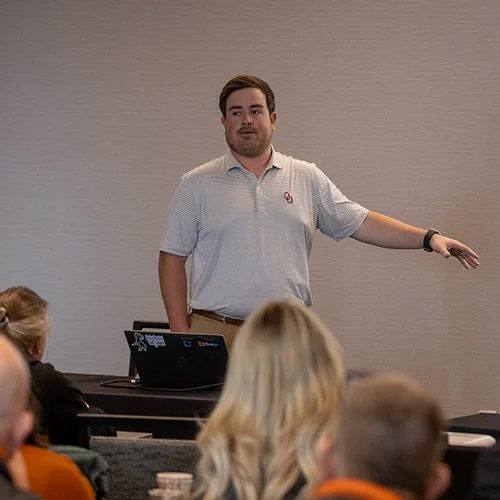 A person in a white polo shirt is giving a presentation in front of an audience. The person is gesturing with their right hand while speaking, and there is a laptop on a table nearby.