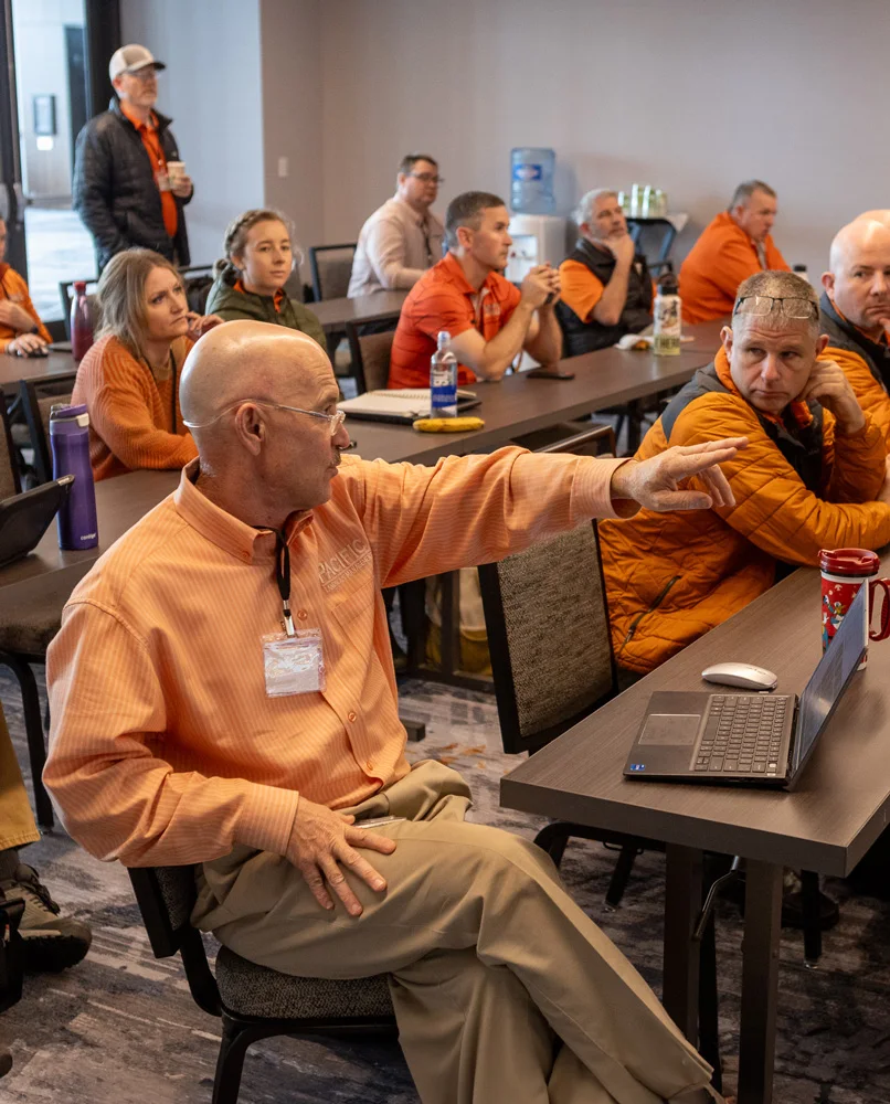 A group of people in an office setting, most wearing orange shirts, are sitting and listening to a speaker. The speaker in the foreground is gesturing towards a laptop on a table.