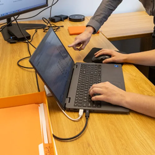 Person typing on a laptop while another person points at the screen in a modern office setting. There is an external monitor and orange office supplies on the wooden desk.
