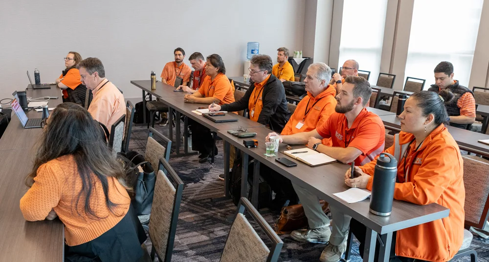 A group of people in orange and black shirts sitting at tables in a conference room, attentively listening to a presentation.