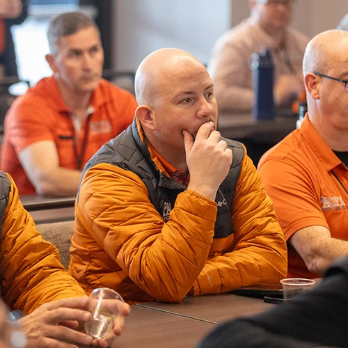 A group of people in a meeting room wearing orange shirts and jackets, with one bald man in the foreground resting his hand on his chin and appearing focused.