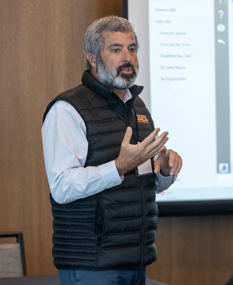 A man with a gray beard is giving a presentation. He is wearing a black puffer vest over a white shirt. A projector screen with indistinct content is visible in the background.