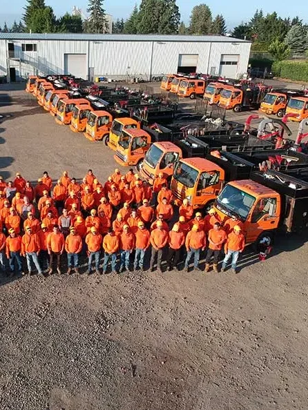 A large group of workers wearing orange safety vests and helmets posing in front of many orange and black utility vehicles in an industrial parking lot with buildings and trees in the background.