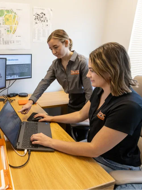 Two women work at a desk with two monitors, a laptop, a folder, and paper documents, inside an office. One woman points at the laptop screen while the other types on the keyboard.