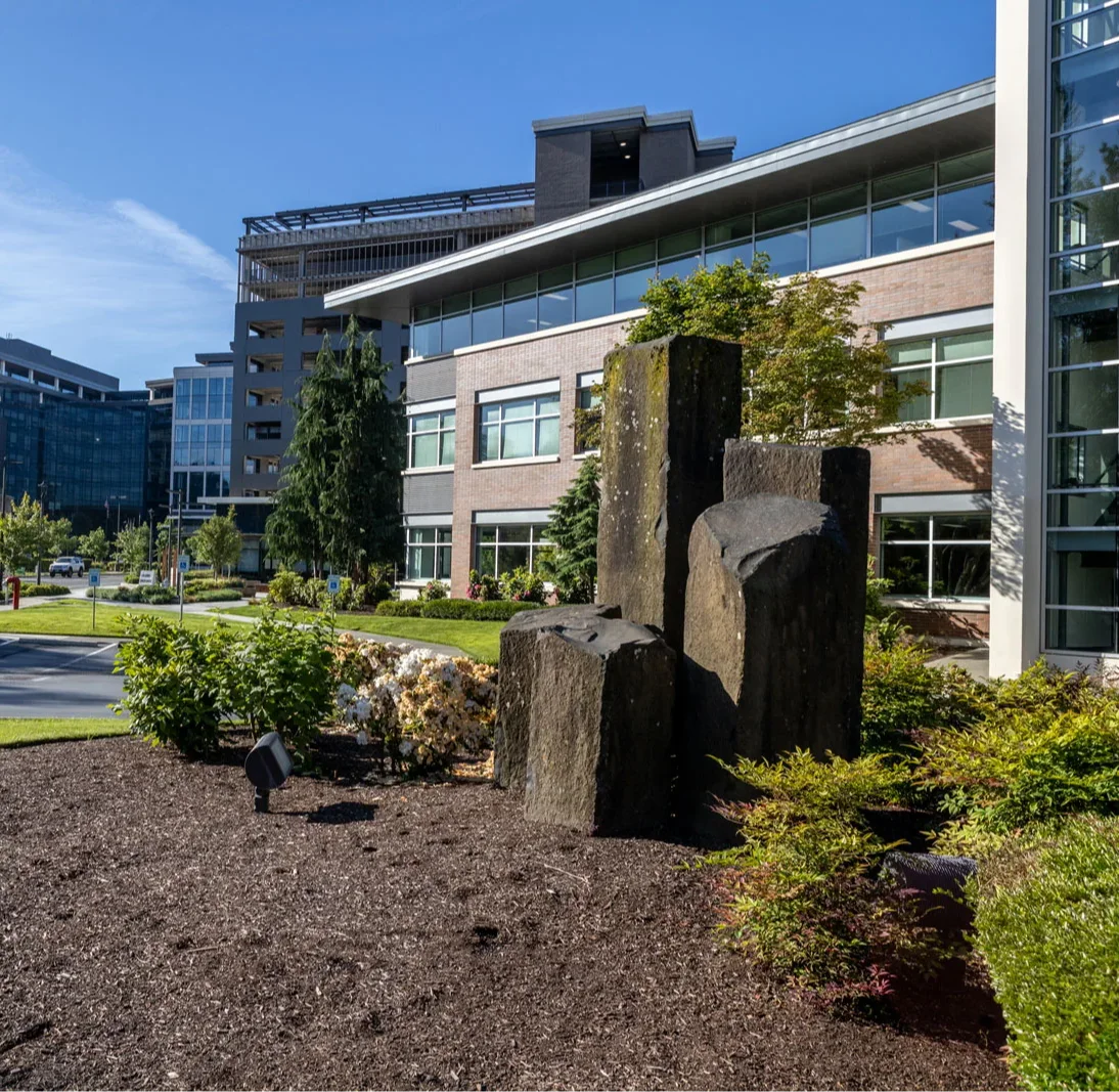 Modern office buildings with large glass windows, surrounded by landscaped greenery and a rock sculpture in the foreground. A clear, blue sky is visible overhead.