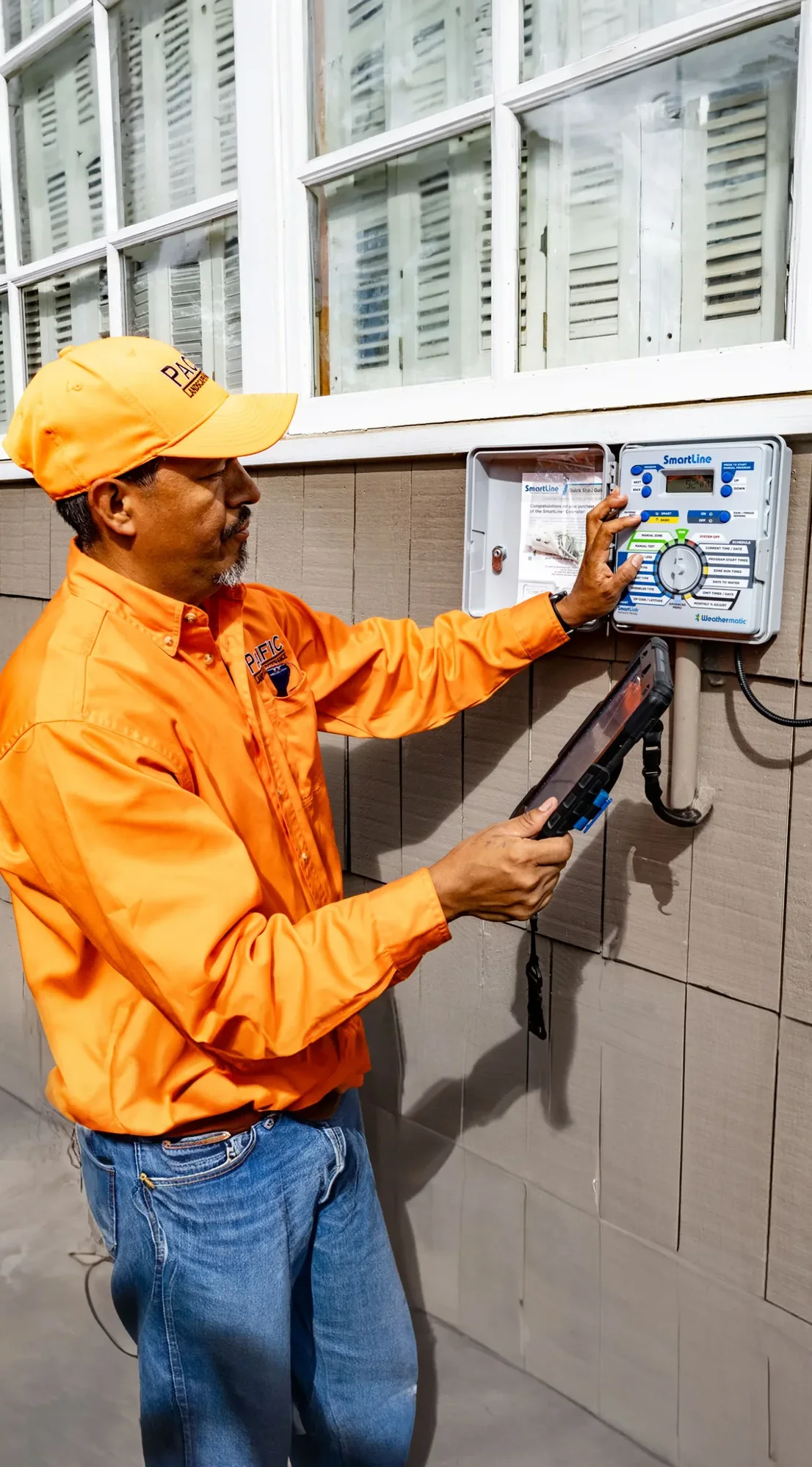 A man in an orange shirt and cap is adjusting settings on an irrigation control panel mounted on a house exterior, with a tablet in his other hand.