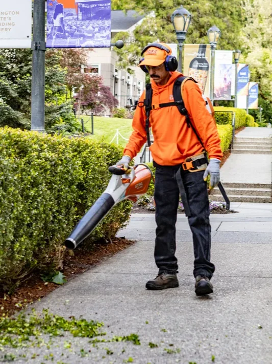 A worker in orange safety gear uses a leaf blower on a sidewalk surrounded by greenery, with a sign in the background promoting a summer concert series.