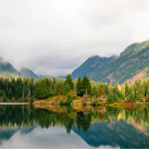 A serene mountain landscape with a forested island reflected in a calm lake. The sky is overcast with low clouds partially covering the mountain peaks in the background.