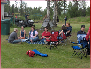 People sit on chairs and a blanket in a grassy area surrounded by trees, with a volleyball game in the background. Some are in conversation, and one wears a distinctive red and black cap.