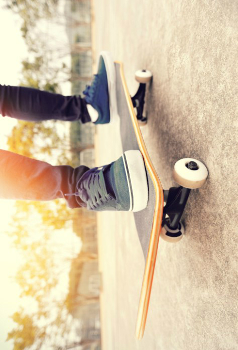 Close-up of a person wearing blue sneakers, standing on a skateboard with one foot back, ready to ride on a smooth concrete surface. Sunlight filters through trees in the background.