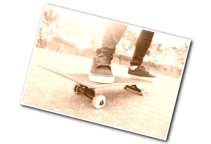 Close-up sepia image of a person standing on a skateboard, wearing dark pants and sneakers, taken at an angle, with trees blurred in the background.