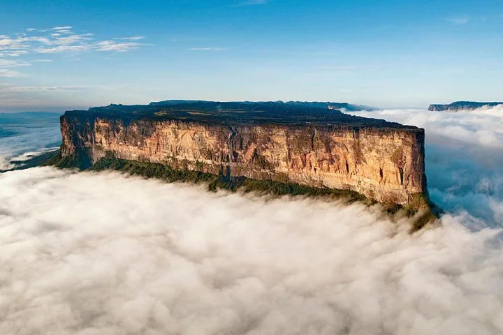 Aerial view of Mount Roraima, a flat-topped mountain surrounded by clouds, with sheer cliffs and green vegetation on top, under a clear blue sky.