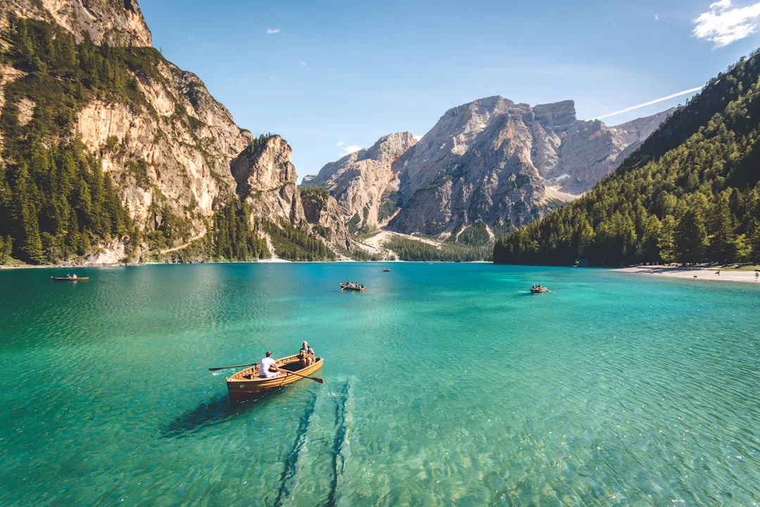A serene lake surrounded by tall mountains and forested hills. Several wooden boats with people paddling are scattered across the turquoise water under a clear blue sky.