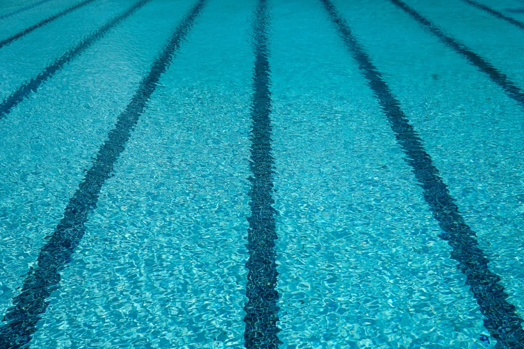 Clear swimming pool water with ripple patterns, showing five dark blue lane markers extending into the distance, indicating lanes for swimmers.